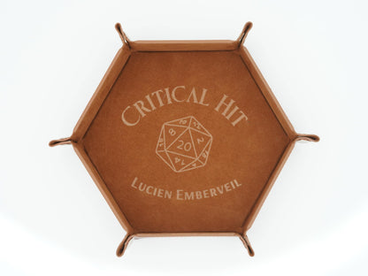 A hexagonal brown leather tray with metal corner supports, featuring an engraved design on the top that reads 'CRITICAL HIT' above a 20-sided die icon and 'LUCIEN EMBERVEIL' below, shown on a white background.