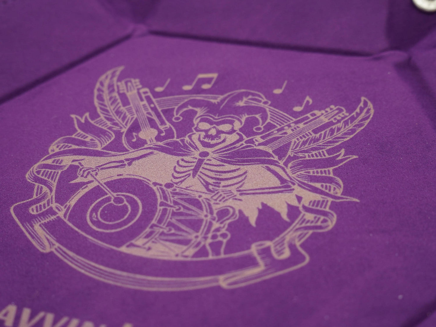 A close-up of a purple velvet or suede-like surface with a light beige engraved illustration of a skeleton playing musical instruments, including a drum and guitar, with decorative banners and notes.