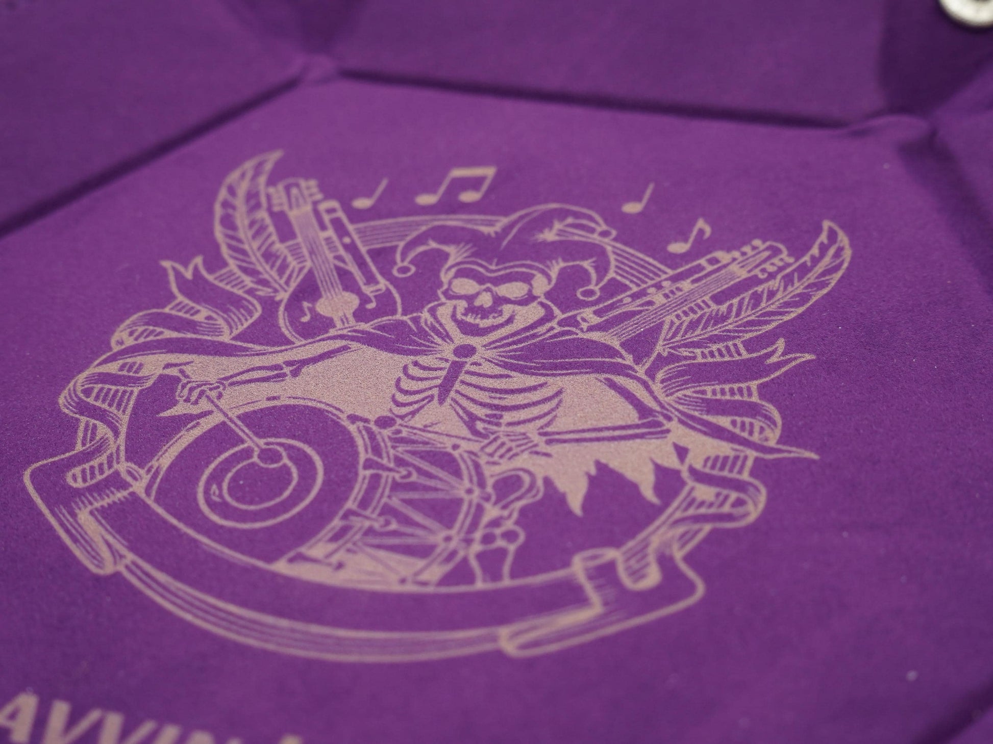 A close-up of a purple velvet or suede-like surface with a light beige engraved illustration of a skeleton playing musical instruments, including a drum and guitar, with decorative banners and notes.