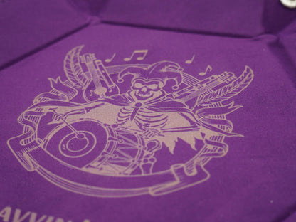 A close-up of a purple velvet or suede-like surface with a light beige engraved illustration of a skeleton playing musical instruments, including a drum and guitar, with decorative banners and notes.