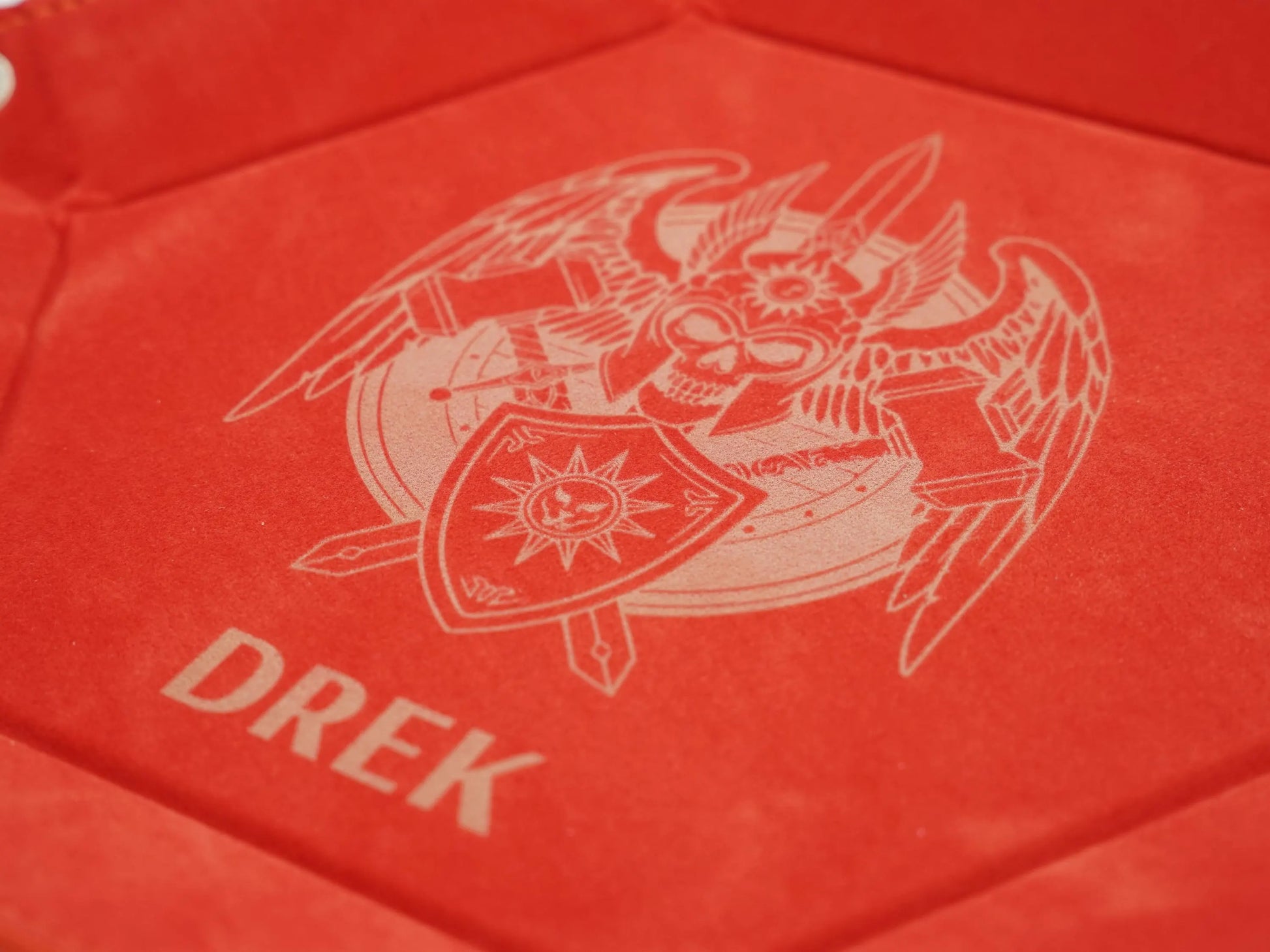 A close-up of a red textured surface featuring a light beige printed emblem of a horned skull with a shield and the word 'DREK' below it.